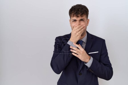 Foto de Joven hombre de negocios hispano usando traje y corbata oliendo algo apestoso y asqueroso, olor intolerable, conteniendo la respiración con los dedos en la nariz. mal olor - Imagen libre de derechos