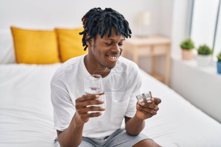Téléchargez les photos : Afro-Américain prenant des pilules assis sur le lit dans la chambre - en image libre de droit