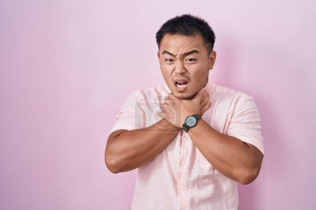 Foto de Joven chino de pie sobre fondo rosa gritando sofocado porque estrangulamiento doloroso. problema de salud. concepto de asfixia y suicidio. - Imagen libre de derechos