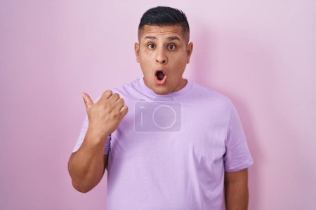 Photo for Young hispanic man standing over pink background surprised pointing with hand finger to the side, open mouth amazed expression. - Royalty Free Image