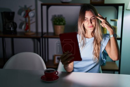 Photo for Young hispanic woman using touchpad sitting on the table at night shooting and killing oneself pointing hand and fingers to head like gun, suicide gesture. - Royalty Free Image