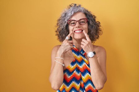 Foto de Mujer de mediana edad con el pelo gris de pie sobre fondo amarillo sonriendo con la boca abierta, los dedos señalando y forzando sonrisa alegre - Imagen libre de derechos