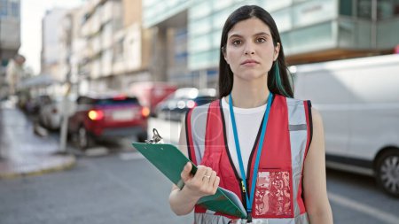 Photo for Young beautiful hispanic woman survey interviewer holding clipboard standing with serious expression at street - Royalty Free Image