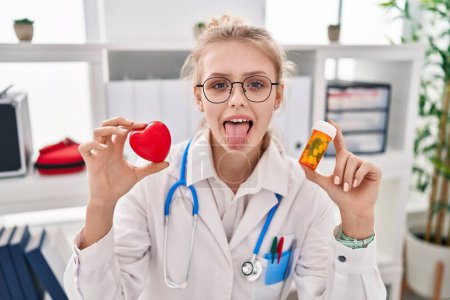 Téléchargez les photos : Jeune médecin caucasien femme tenant des pilules et coeur langue collante heureux avec une expression drôle. - en image libre de droit