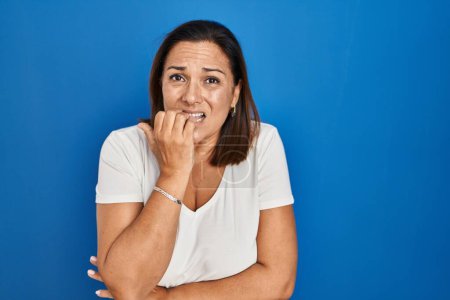 Téléchargez les photos : Femme mûre hispanique debout sur fond bleu regardant stressé et nerveux avec les mains sur les ongles mordants de la bouche. problème d'anxiété. - en image libre de droit