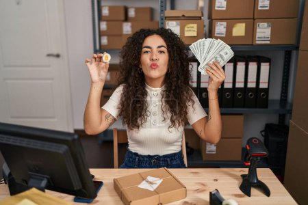 Photo for Young hispanic woman working at small business ecommerce holding money and bitcoin looking at the camera blowing a kiss being lovely and sexy. love expression. - Royalty Free Image