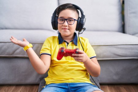 Foto de Joven niño hispano jugando videojuego sosteniendo controlador usando auriculares sonriendo alegre presentando y señalando con la palma de la mano mirando a la cámara. - Imagen libre de derechos