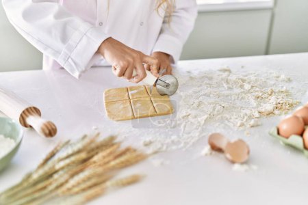 Foto de Young woman wearing cook uniform cutting pasta dough at kitchen - Imagen libre de derechos