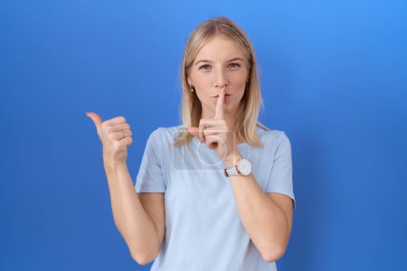 Téléchargez les photos : Jeune femme caucasienne portant un t-shirt bleu décontracté demandant à être calme avec le doigt sur les lèvres pointant avec la main sur le côté. silence et concept secret. - en image libre de droit