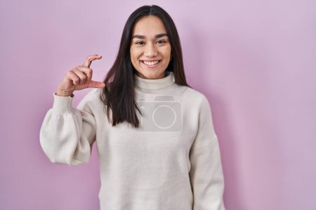 Téléchargez les photos : Jeune femme sud-asiatique debout sur fond rose geste souriant et confiant avec la main faisant signe de petite taille avec les doigts à la recherche et la caméra. concept de mesure. - en image libre de droit