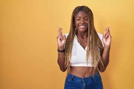 Téléchargez les photos : Femme afro-américaine avec les cheveux tressés debout sur fond jaune geste doigt croisé souriant avec espoir et les yeux fermés. chance et concept superstitieux. - en image libre de droit