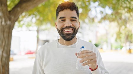 Foto de Joven hombre hispano sonriendo confiado sosteniendo botella de agua en el parque - Imagen libre de derechos