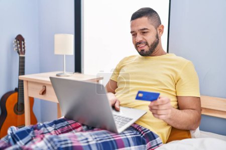Téléchargez les photos : Jeune homme latin utilisant ordinateur portable et carte de crédit assis sur le lit dans la chambre - en image libre de droit
