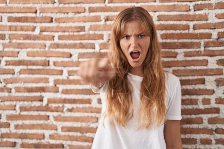 Foto de Mujer joven caucásica de pie sobre la pared de ladrillos señalando disgustado y frustrado a la cámara, enojado y furioso con usted - Imagen libre de derechos