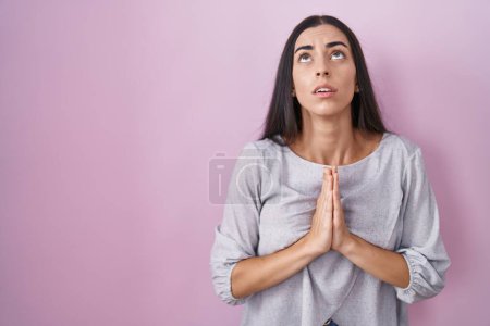 Téléchargez les photos : Jeune femme brune debout sur fond rose mendiant et priant avec les mains avec l'expression d'espoir sur le visage très émotionnel et inquiet. mendier. - en image libre de droit