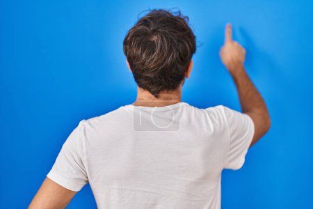 Photo for Hispanic young man standing over blue background posing backwards pointing ahead with finger hand - Royalty Free Image