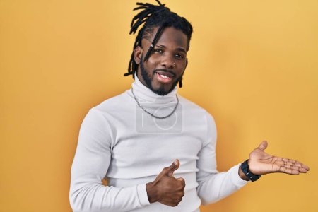Photo for African man with dreadlocks wearing turtleneck sweater over yellow background showing palm hand and doing ok gesture with thumbs up, smiling happy and cheerful - Royalty Free Image