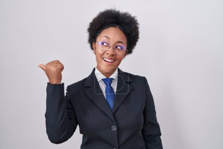 Foto de Hermosa mujer africana con el pelo rizado usando chaqueta de negocios y gafas sonriendo con la cara feliz mirando y señalando a un lado con el pulgar hacia arriba. - Imagen libre de derechos