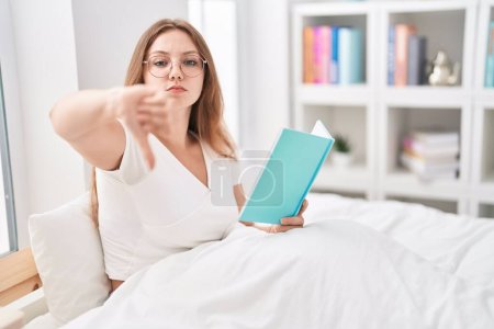 Téléchargez les photos : Jeune femme caucasienne assise sur le lit à la maison lisant un livre avec un visage en colère, signe négatif montrant dégoût avec les pouces vers le bas, concept de rejet - en image libre de droit