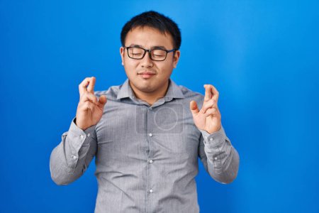 Téléchargez les photos : Jeune chinois debout sur fond bleu geste doigt croisé souriant avec espoir et les yeux fermés. chance et concept superstitieux. - en image libre de droit