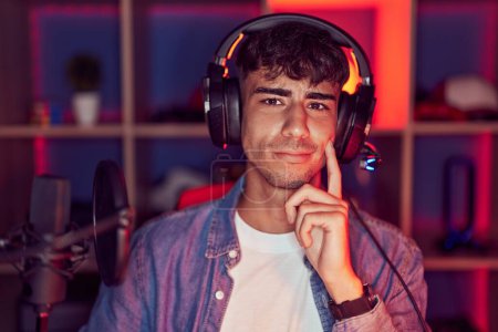 Téléchargez les photos : Jeune homme hispanique jouant à des jeux vidéo pensant concentré sur le doute avec le doigt sur le menton et regardant vers le haut se demandant - en image libre de droit