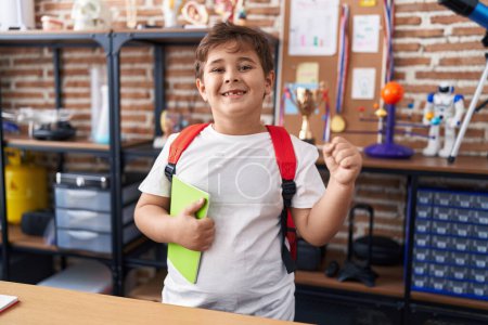 Photo for Little hispanic boy wearing student backpack and holding book at school class screaming proud, celebrating victory and success very excited with raised arm - Royalty Free Image