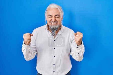 Photo for Middle age man with grey hair standing over blue background excited for success with arms raised and eyes closed celebrating victory smiling. winner concept. - Royalty Free Image