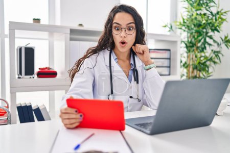 Foto de Young doctor woman working on online appointment scared and amazed with open mouth for surprise, disbelief face - Imagen libre de derechos