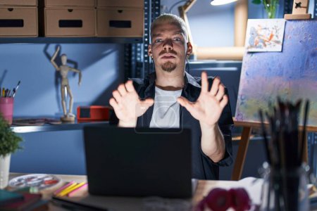 Téléchargez les photos : Jeune homme caucasien utilisant un ordinateur portable la nuit au studio d'art effrayé et terrifié par l'expression de la peur arrêter geste avec les mains, criant en état de choc. concept de panique. - en image libre de droit