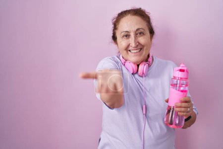 Foto de Mujer mayor que usa ropa deportiva y auriculares sonriendo alegre ofreciendo la palma de la mano dando asistencia y aceptación. - Imagen libre de derechos