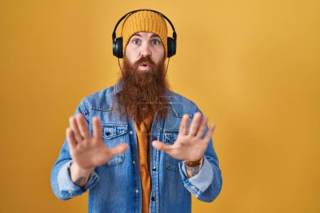 Photo for Caucasian man with long beard listening to music using headphones moving away hands palms showing refusal and denial with afraid and disgusting expression. stop and forbidden. - Royalty Free Image