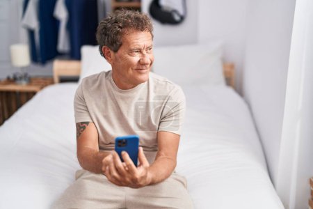 Photo for Middle age man using smartphone sitting on bed at bedroom - Royalty Free Image