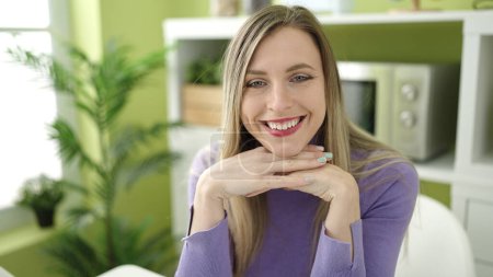 Téléchargez les photos : Jeune femme blonde souriante confiante assise sur la table à la maison - en image libre de droit