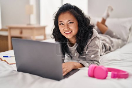 Téléchargez les photos : Jeune femme chinoise utilisant un ordinateur portable couché sur le lit dans la chambre - en image libre de droit