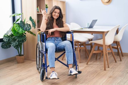 Téléchargez les photos : Jeune adolescente assise en fauteuil roulant au salon souriant heureux pointant avec la main et le doigt sur le côté - en image libre de droit