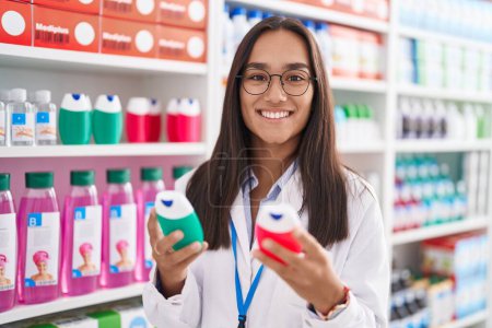 Foto de Young beautiful hispanic woman pharmacist holding toothpaste bottles at pharmacy - Imagen libre de derechos