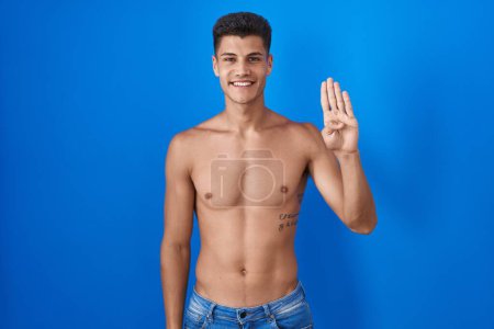 Foto de Joven hombre hispano de pie sin camisa sobre fondo azul mostrando y señalando con los dedos número cuatro mientras sonríe confiado y feliz. - Imagen libre de derechos
