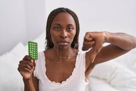 Téléchargez les photos : Femme afro-américaine tenant des pilules contraceptives avec le visage en colère, signe négatif montrant aversion avec les pouces vers le bas, concept de rejet - en image libre de droit