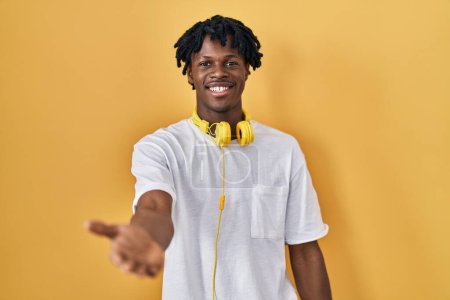 Téléchargez les photos : Jeune homme africain avec dreadlocks debout sur fond jaune souriant joyeuse offrant main de paume donnant assistance et acceptation. - en image libre de droit