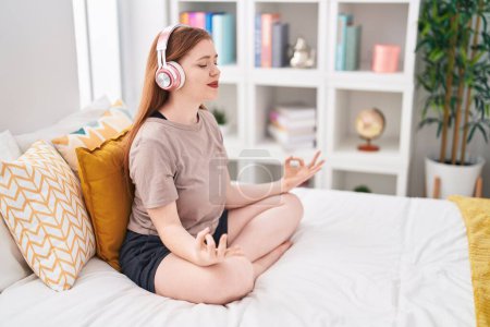 Young redhead woman doing yoga exercise sitting on bed at bedroom