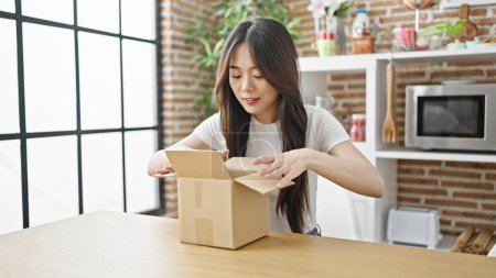 Photo for Young chinese woman unpacking cardboard box sitting on table with serious expression at dinning room - Royalty Free Image