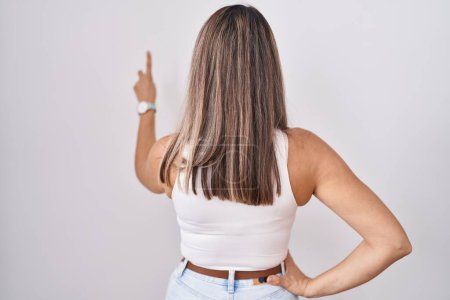Foto de Mujer joven hispana de pie sobre fondo blanco posando hacia atrás apuntando hacia adelante con la mano del dedo - Imagen libre de derechos