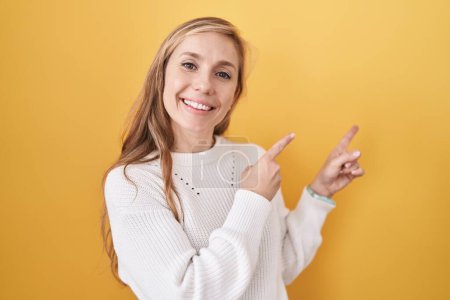 Foto de Mujer joven caucásica vistiendo suéter blanco sobre fondo amarillo sonriendo y mirando a la cámara apuntando con dos manos y dedos hacia un lado. - Imagen libre de derechos