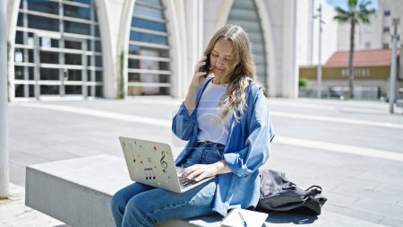 Foto de Joven estudiante rubia usando laptop hablando en smartphone en la universidad - Imagen libre de derechos