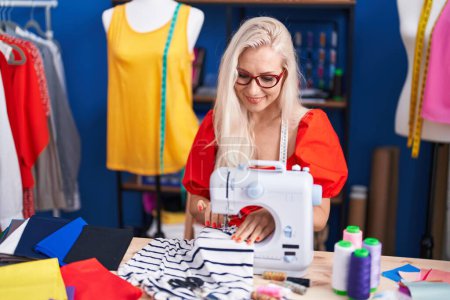 Téléchargez les photos : Jeune femme blonde tailleur souriant confiant en utilisant une machine à coudre au studio de couture - en image libre de droit
