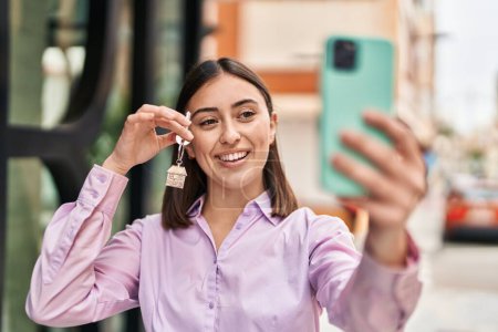 Téléchargez les photos : Jeune femme hispanique faire selfie par le smartphone tenant la clé de la nouvelle maison à la rue - en image libre de droit