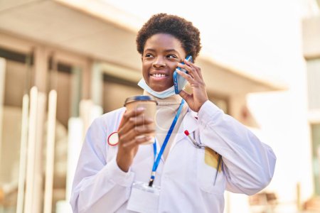 Téléchargez les photos : Femme afro-américaine portant un uniforme de médecin et un masque médical parlant sur le smartphone buvant du café dans la rue - en image libre de droit