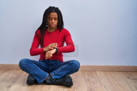 Photo for Young african american with braids sitting on the floor at home checking the time on wrist watch, relaxed and confident - Royalty Free Image
