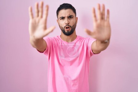 Foto de Joven hispano parado sobre fondo rosa haciendo gesto de stop con palmas de las manos, expresión de enojo y frustración - Imagen libre de derechos