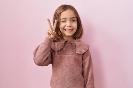 Photo for Little hispanic girl standing over pink background smiling with happy face winking at the camera doing victory sign. number two. - Royalty Free Image